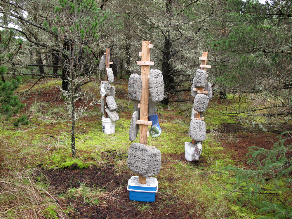 a trio of sculptures comprised of vertical wooden suppoerts grounded in concrete filled buckets, hung with concrete slabs and concrete filled gloves and shoes